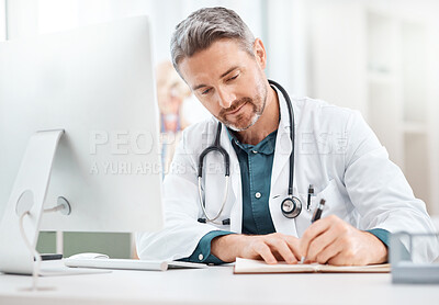 Buy stock photo Shot of a mature doctor writing notes while working on a computer in a medical office