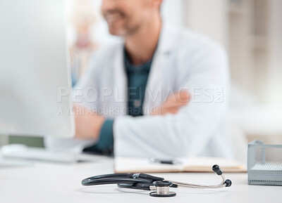 Buy stock photo Closeup shot of a stethoscope on a table with a doctor working on a computer in the background in a medical office