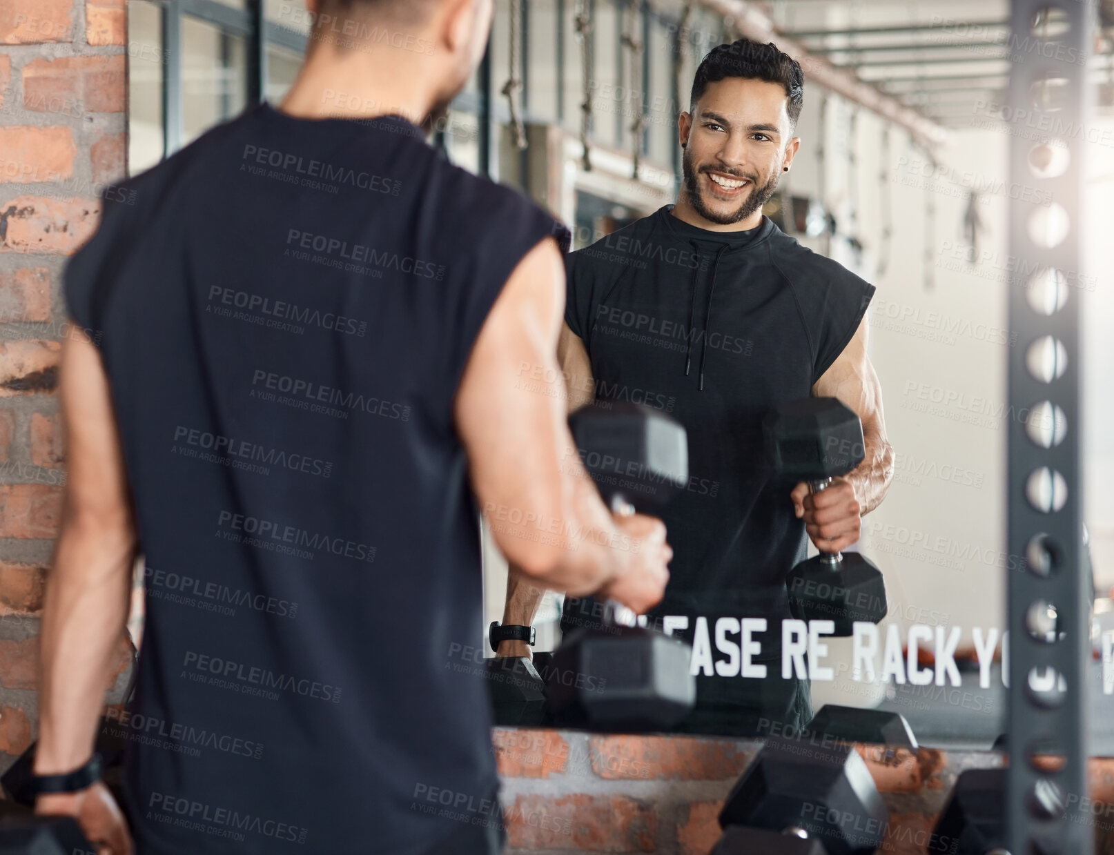 Buy stock photo Shot of a sporty young man looking at himself in a mirror while exercising with dumbbells in a gym