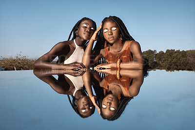 Buy stock photo Shot of two attractive young women sitting next to a pond with their reflection in it in nature