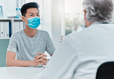 Buy stock photo Shot of a young man wearing a face mask and sitting with his doctor during his consultation in the clinic