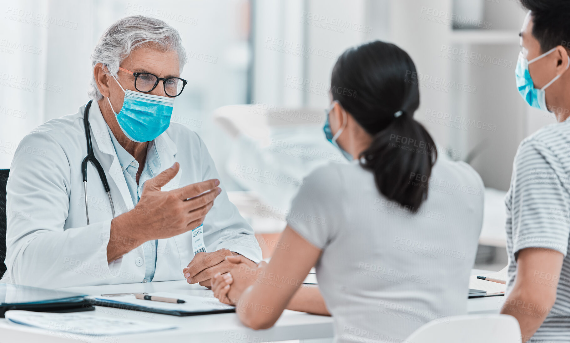Buy stock photo Shot of a mature doctor wearing a face mask and sitting with his patients during a consultation in his clinic