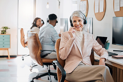 Buy stock photo Shot of a mature businesswoman sitting in the office and wearing a headset while her colleagues work behind her