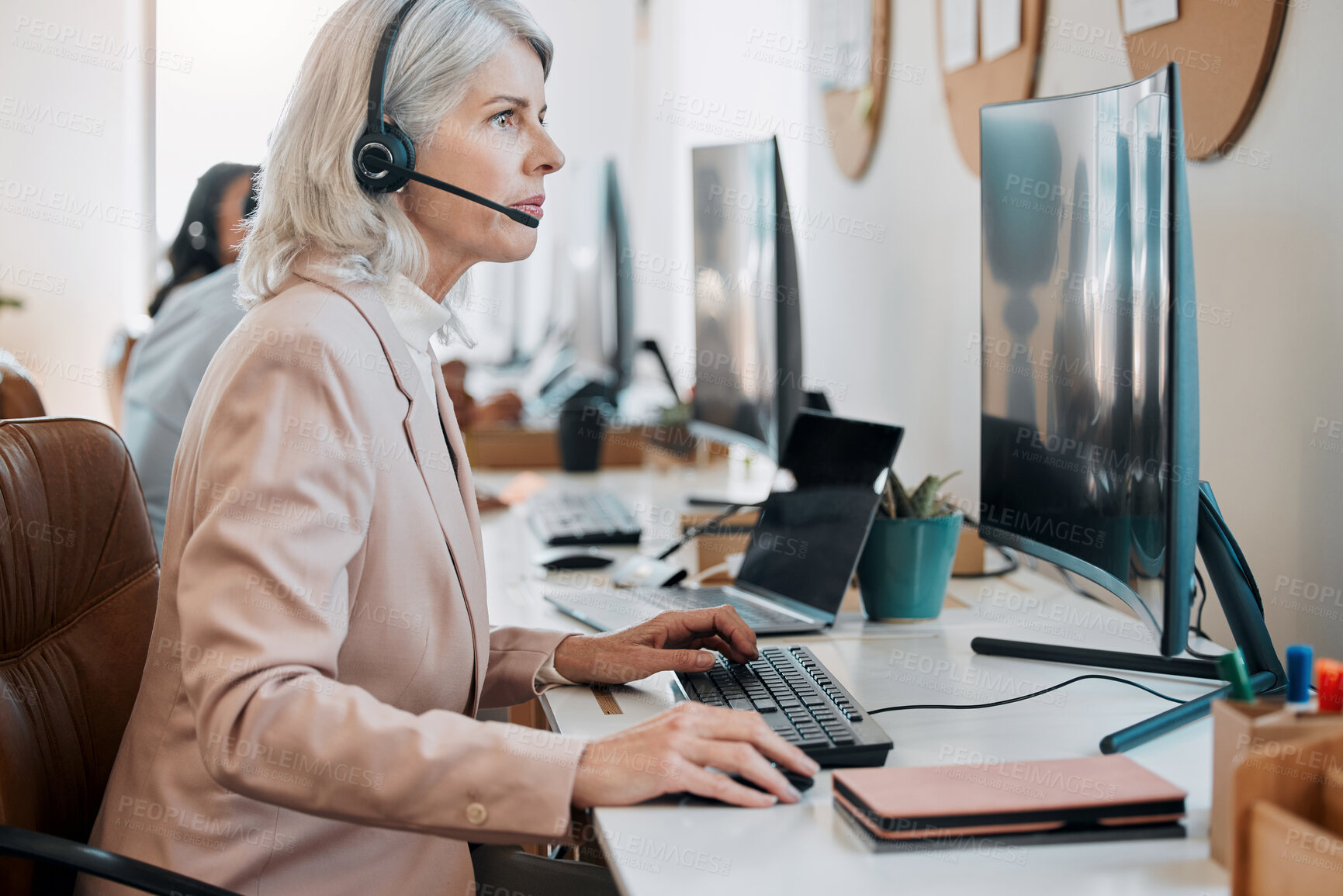 Buy stock photo Shot of a mature agent sitting in the office and using her computer while her colleagues work behind her