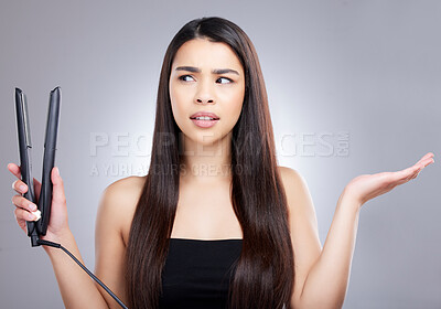 Buy stock photo Studio shot of an attractive young woman looking confused while using a flat iron to straighten her hair against a grey background