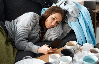 Buy stock photo Shot of a young woman lying on her messy couch looking depressed and using her smartphone
