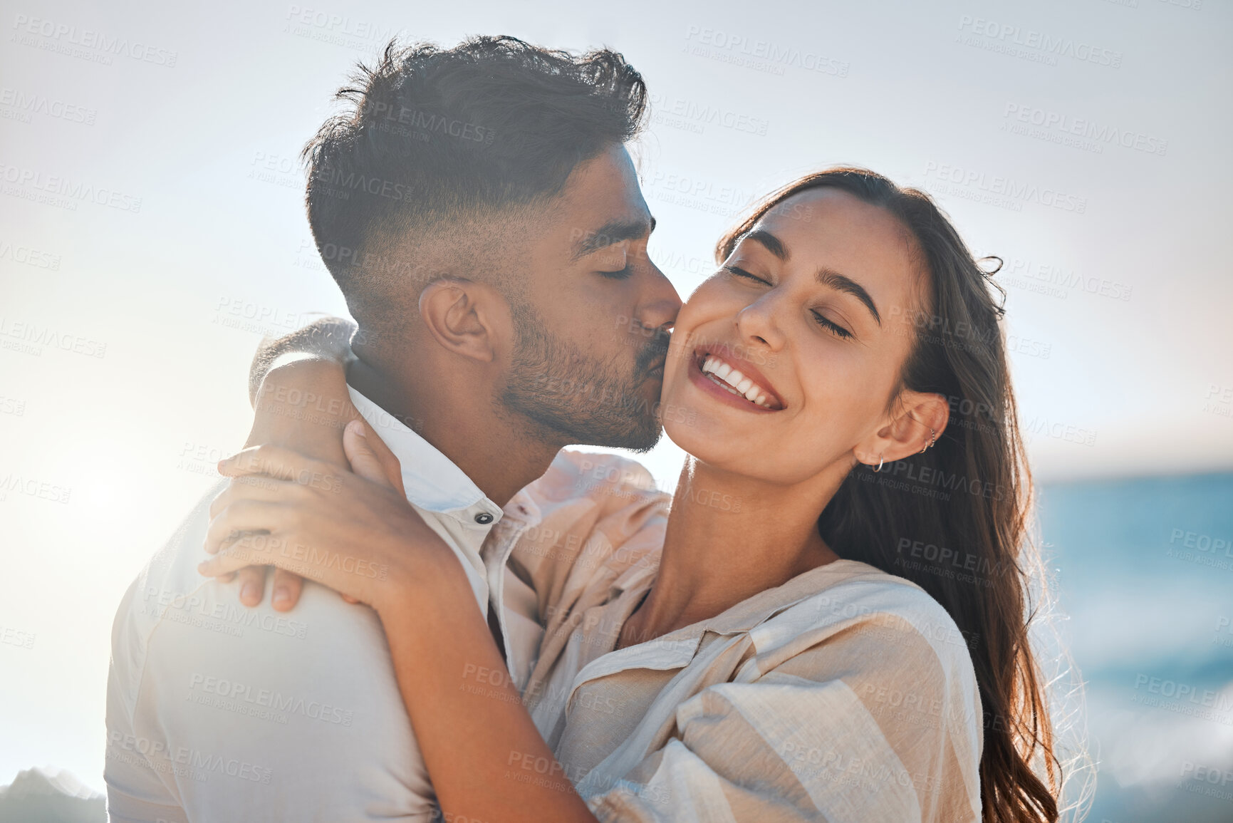 Buy stock photo Interracial, kiss or couple hug at ocean for love on vacation, valentines day or holiday in Spain. Happy, relationship and people at sea for bonding together for romantic trip at beach with smile