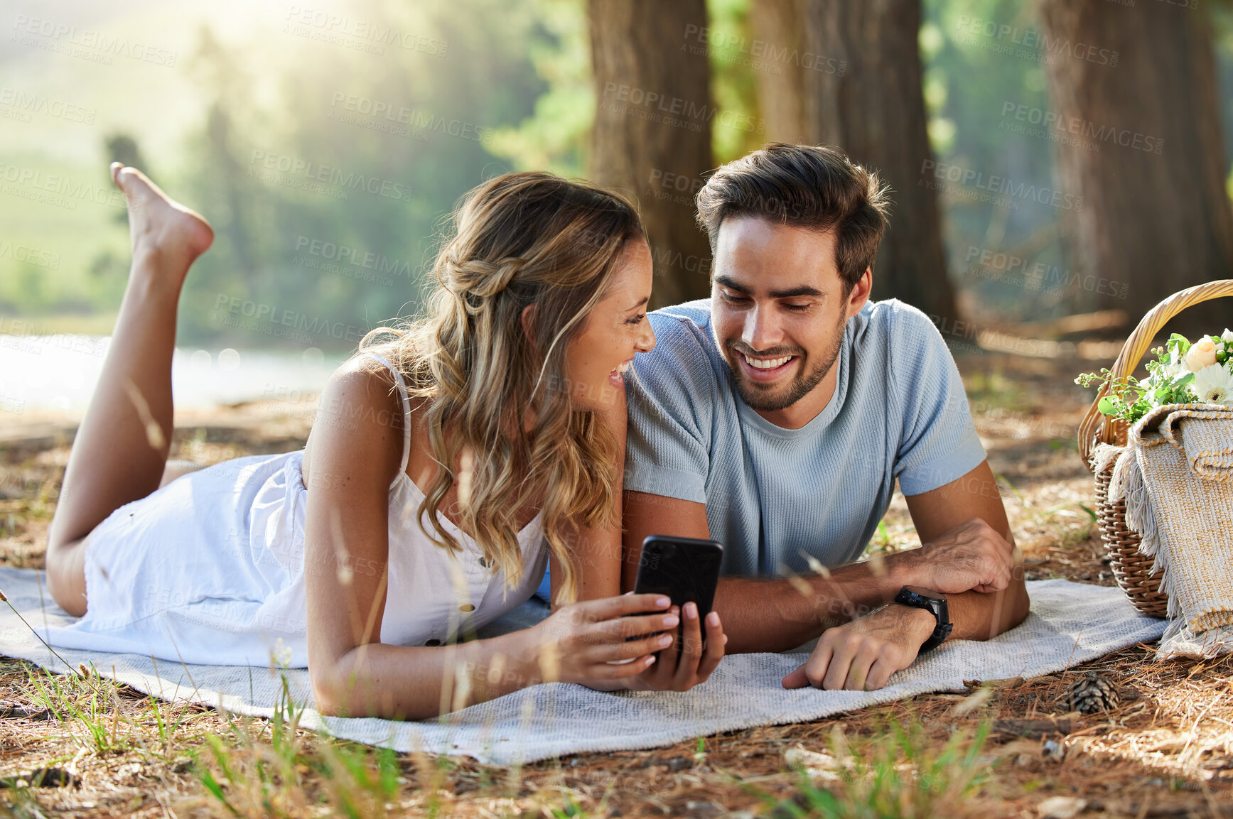 Buy stock photo Couple on picnic with smartphone, relax together in nature and social media with travel and bonding outdoor. Happiness, man and woman lying down and scroll internet in forest with smile