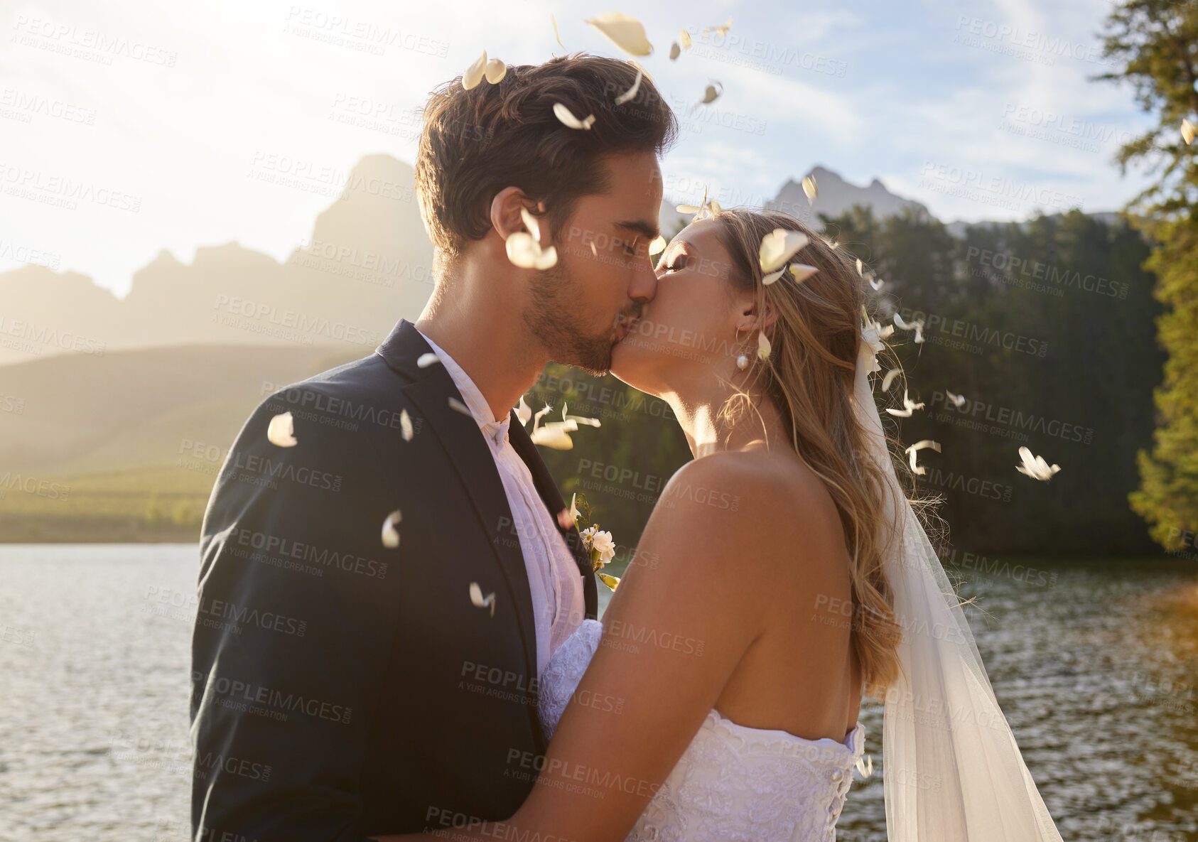 Buy stock photo Love, wedding and confetti with a couple kissing by a lake outdoor in celebration of a marriage for romance. Water, summer or kiss with a bride and groom bonding together in tradition after ceremony