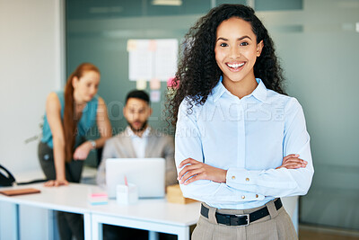 Buy stock photo Shot of an attractive young businesswoman standing in the office with her arms folded while her colleagues work behind her