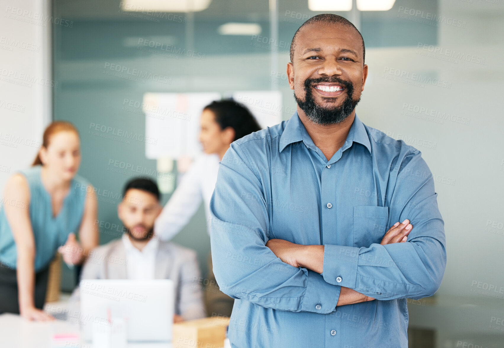 Buy stock photo Shot of a handsome mature businessman standing in the office with his arms folded while his colleagues work behind him