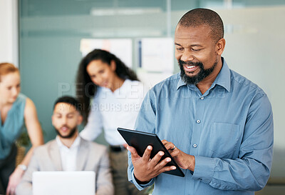 Buy stock photo Shot of a mature businessman standing in the office and using a digital tablet while his colleagues work behind him