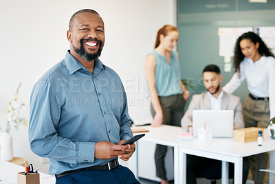 Buy stock photo Shot of a mature businessman sitting in the office and using his cellphone while his colleagues work behind him
