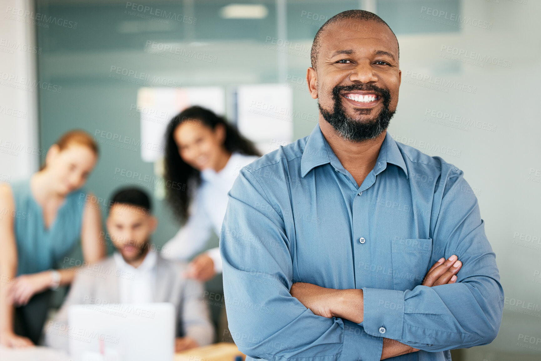 Buy stock photo Shot of a handsome mature businessman standing in the office with his arms folded while his colleagues work behind him