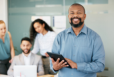 Buy stock photo Shot of a mature businessman standing in the office and using a digital tablet while his colleagues work behind him