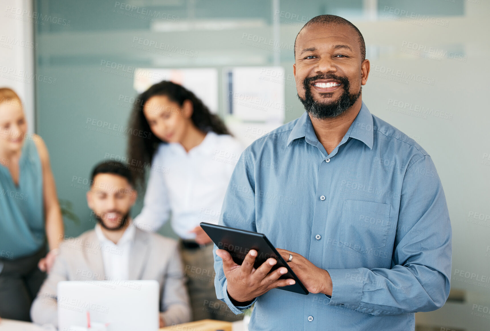 Buy stock photo Shot of a mature businessman standing in the office and using a digital tablet while his colleagues work behind him