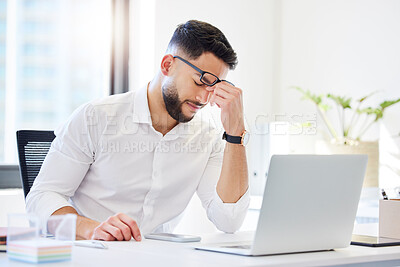 Buy stock photo Shot of a handsome young businessman sitting alone in the office and feeling stressed while using his laptop