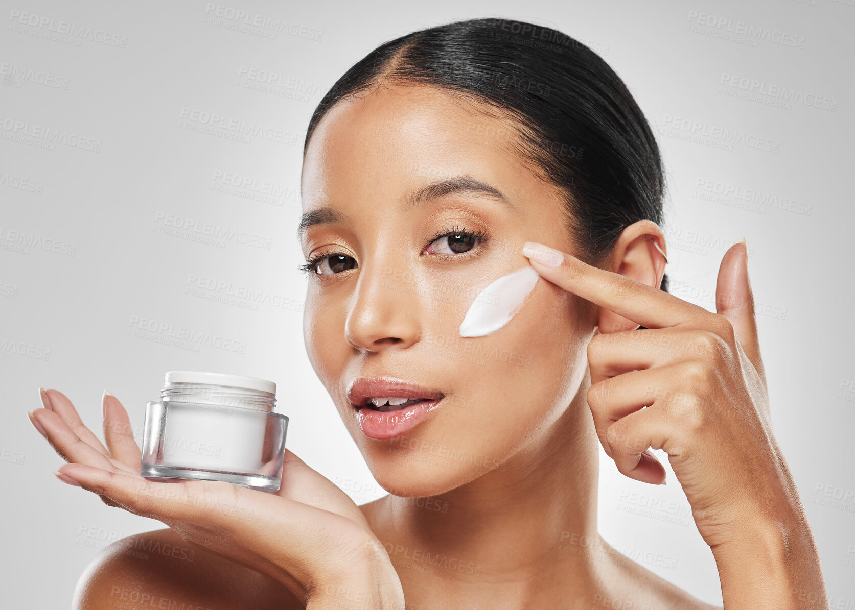 Buy stock photo Studio portrait of an attractive young woman applying moisturiser on her face against a grey background