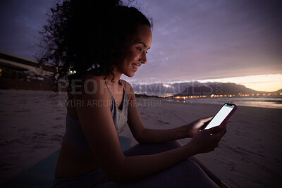 Buy stock photo Shot of a fit young woman using her cellphone while sitting on the beach at night