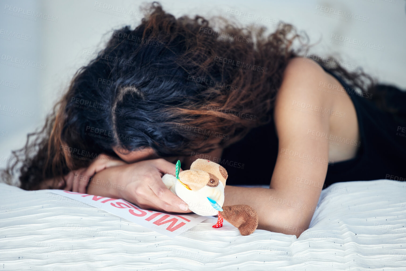 Buy stock photo Shot of a young woman looking sad while holding a teddy bear and lying on a bed alongside a missing person poster