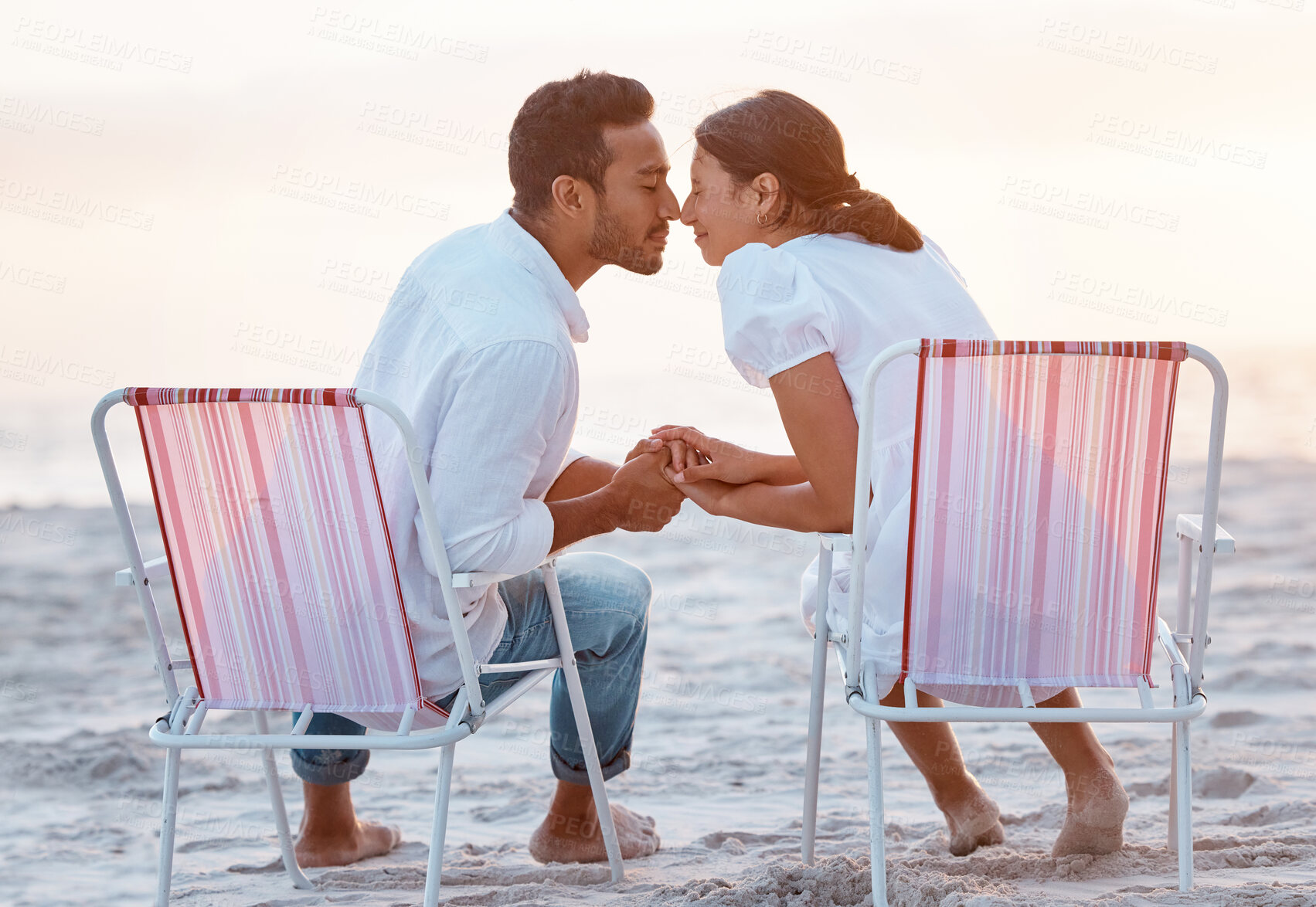 Buy stock photo Shot of a young couple holding hands while sitting on their beach chairs