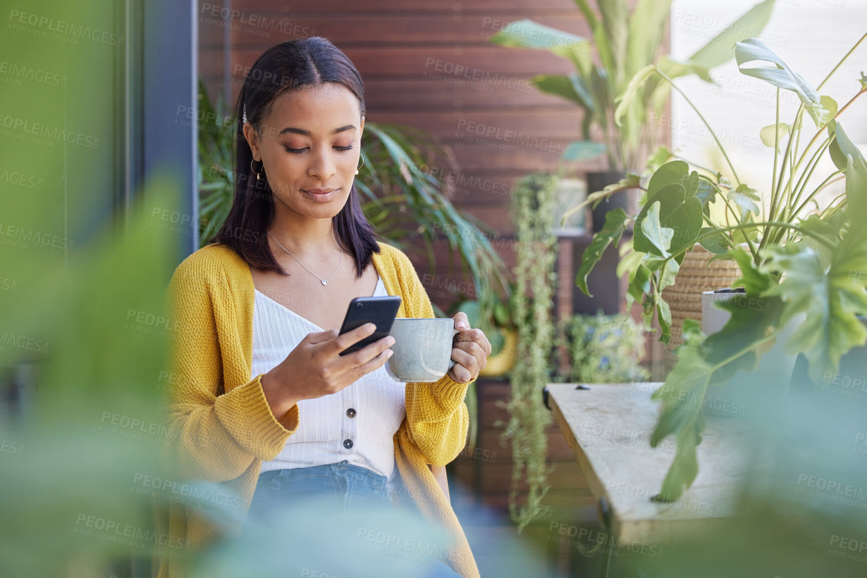 Buy stock photo Shot of a young woman using a phone while drinking coffee on the balcony at home