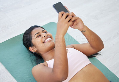 Buy stock photo Shot of a sporty young woman using a cellphone while lying on an exercise mat in a yoga studio