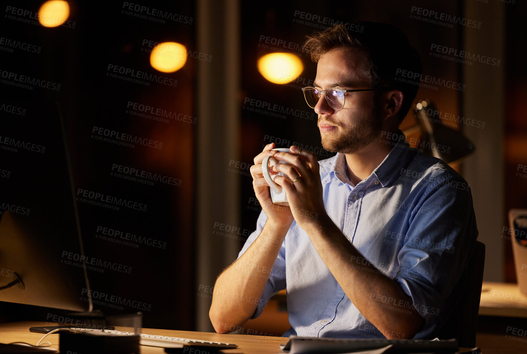 Buy stock photo Shot of a young businessman drinking coffee while working on a computer in an office at night