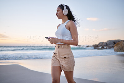 Buy stock photo Shot of a woman wearing headphones and using her cellphone while walking on the beach