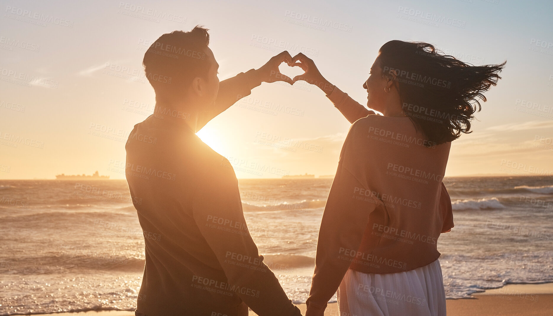 Buy stock photo Couple, sunset and love sign at beach with hands for date, relationship and romantic moment together. Man, woman and heart gesture by ocean with partner and back view for travel, holiday and loyalty