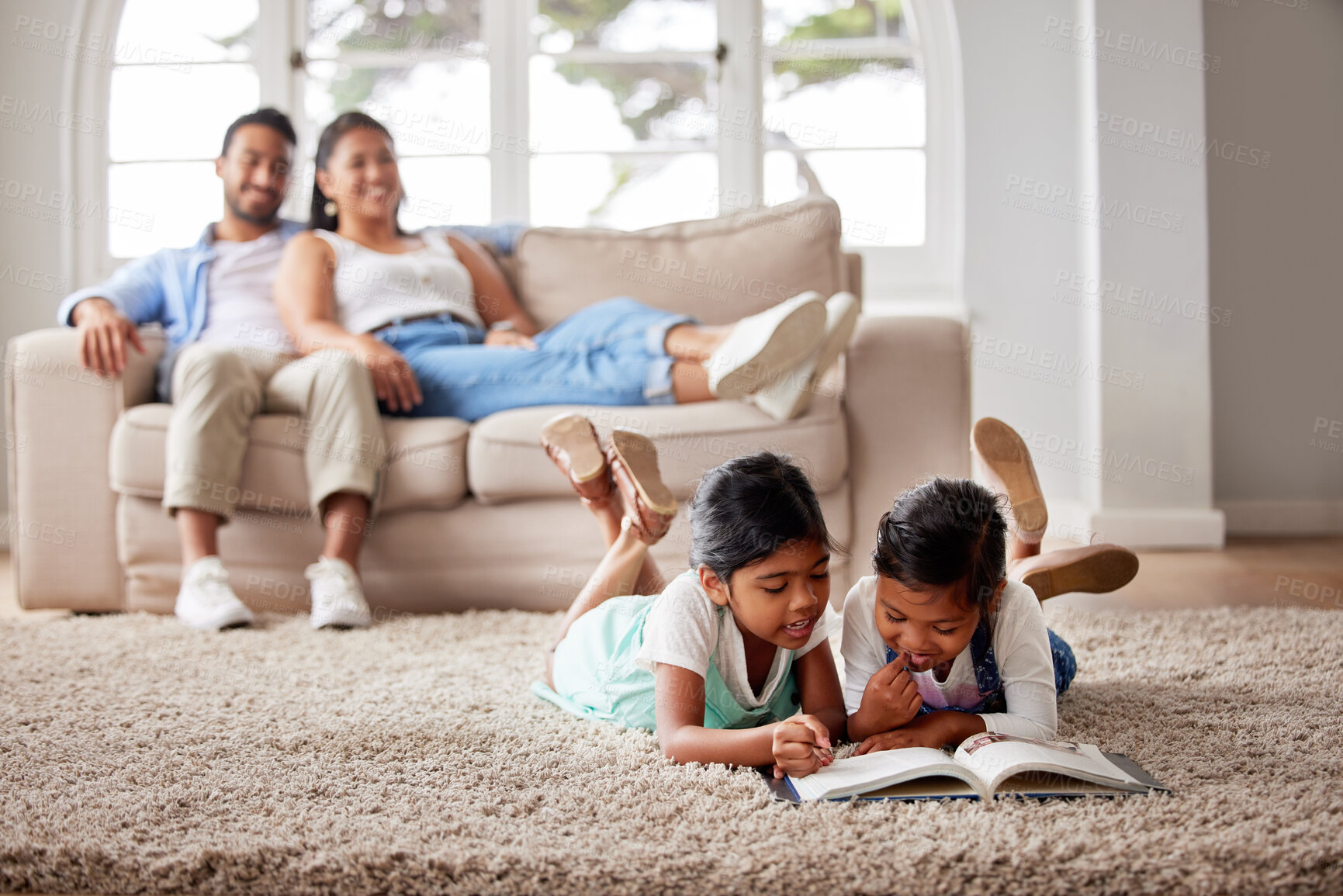 Buy stock photo Two young children reading a book on the living room floor while their parents relax on a sofa in the background at home