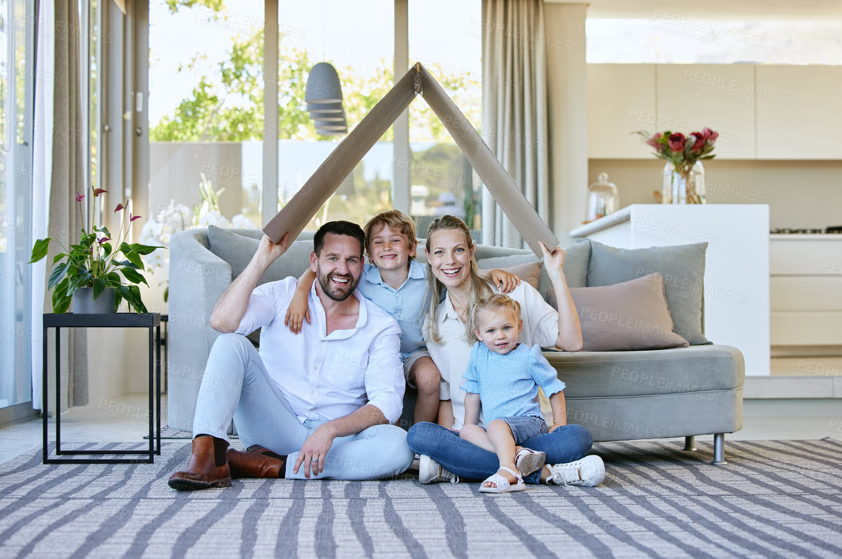 Buy stock photo Full length portrait of an affectionate family of four holding a cardboard roof over their heads while sitting in the living room at home