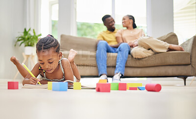 Buy stock photo Shot of an adorable little girl lying on the living room floor and colouring while her parents bond behind her