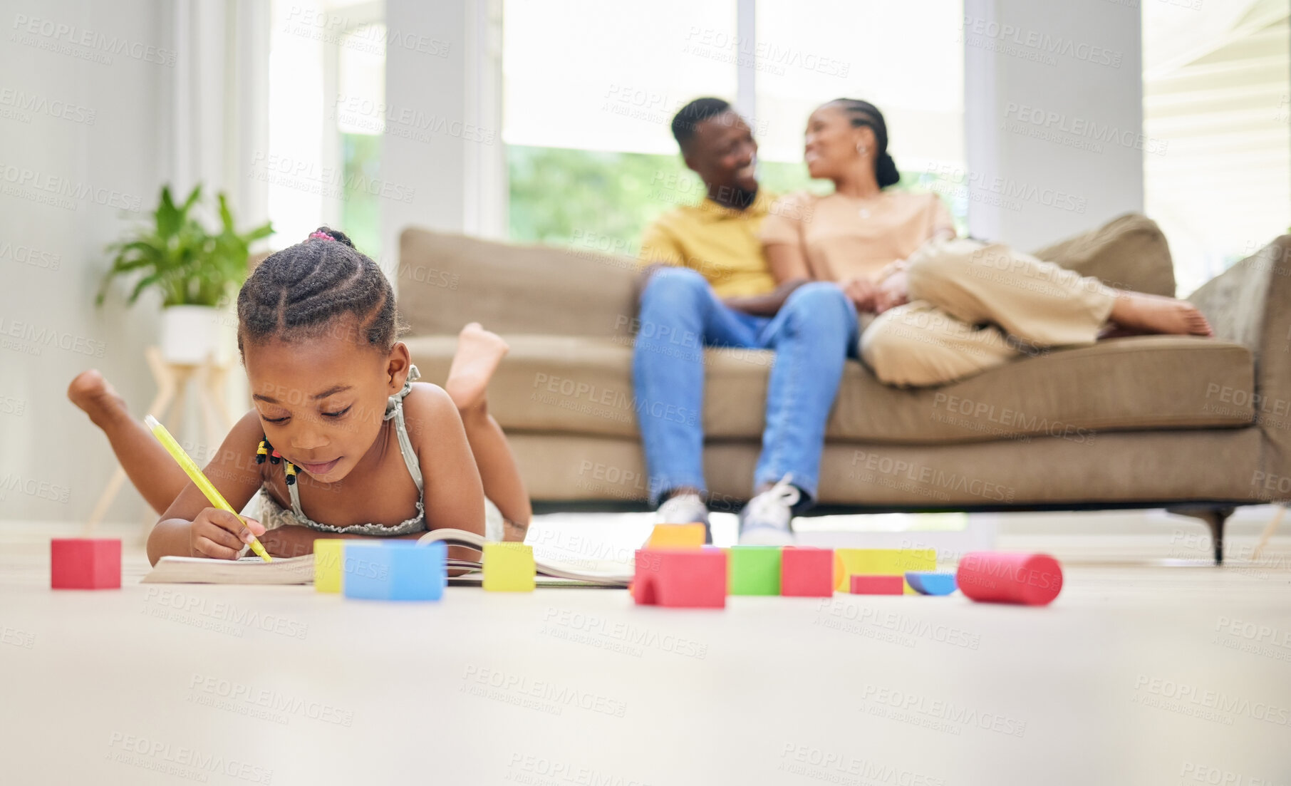 Buy stock photo Shot of an adorable little girl lying on the living room floor and colouring while her parents bond behind her