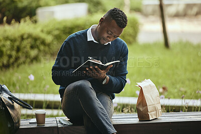 Black businessman writing in a notebook while sitting in a park. Taking a break from the office to do some planning in his diary