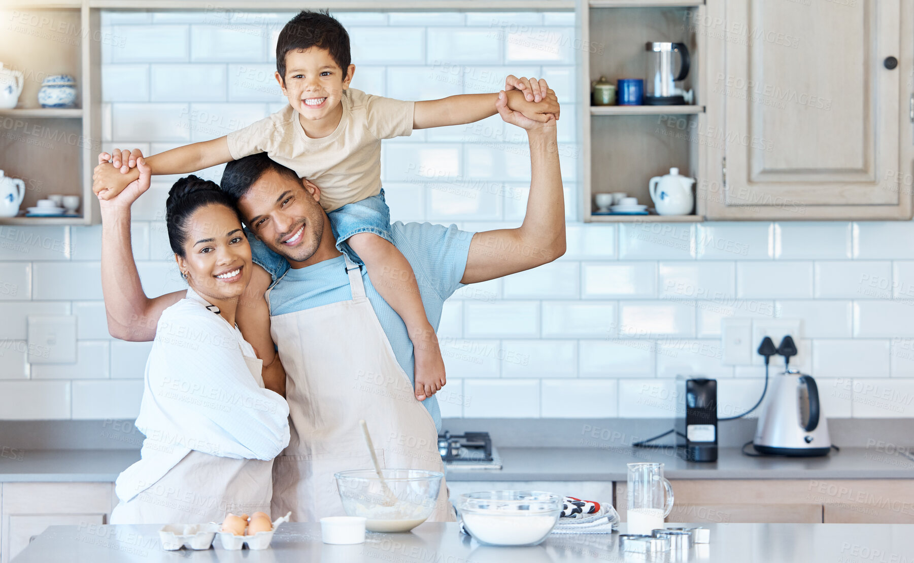 Buy stock photo Portrait of happy young mixed race couple bonding with their adorable son in a home kitchen while baking. Hispanic husband carrying a child on his shoulders while his wife embraces him. Time together