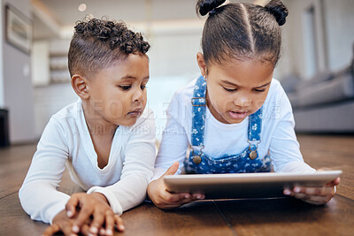 Two young mixed race children using a digital tablet while lying together on the floor at home. Young boy and girl siblings browsing the internet online to play games and learn from educational apps