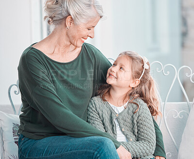Adorable little girl smiling and looking up at her grandmother while sitting together and bonding at home. Beautiful mature caucasian woman showing love and affection to her granddaughter