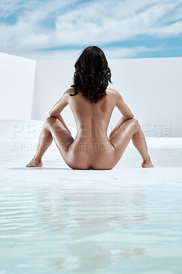 Young mixed race woman posing naked in water while sitting against a blue sky outdoors. Rear view shot of one female posing sensual, sexy and seductive while flaunting her feminine curvy body in nude