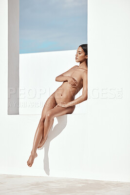 Serene, nude, hispanic model posing against a white background outside. Body of a sensual, beautiful young model. Naked woman with perfect skin covering her chest sitting outside.