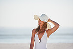 A smiling young woman in a white swimsuit holding her hat and enjoying a day at the beach.A carefree young woman in swimwear thinking and relaxing by the sea. A woman on holiday at the beach