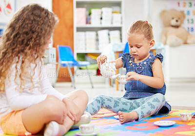 Two little girls having a tea party while playing on a colourful mat on the floor at preschool or kindergarten. Happy caucasian kids engaged in fun creative imaginative learning and development