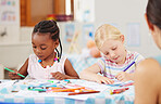 African american girl sitting at a table and colouring at pre-school or kindergarten with her caucasian classmate . Young female children using colourful pencils to draw pictures in a class at school