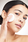 Beautiful young woman doing her skincare routine standing against a grey studio background alone. Female using a cotton pad on her face while standing against a background