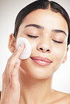 Beautiful young happy woman doing her skincare routine standing against a grey studio background alone. Content female using a cotton pad on her face while standing against a background