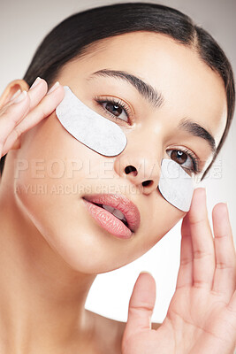 Beautiful young happy woman applying eye patches and doing her skincare routine standing against a grey studio background alone. One content female taking care of her skin while standing against a background