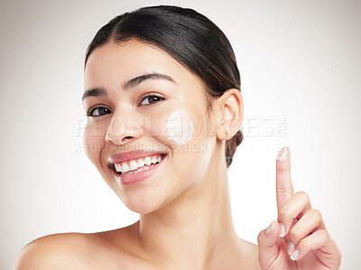Buy stock photo Portrait of a young happy mixed race woman applying lotion to her face while standing against a grey studio background alone. One cheerful hispanic female doing her skincare routine while standing against a background