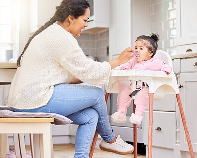 Mixed race single mother feeding her adorable little baby girl in a feeding chair in the kitchen at home. Young hispanic mother bonding with her daughter while eating a healthy, nutritious meal