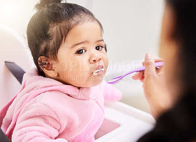 One mixed race baby girl being fed with a spoon by mom at home. Cute toddler with her mouth stuffed with porridge sitting in a feeding chair to enjoy some soft foods. Nutrition for healthy development