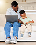 Happy cheerful young african american man using a laptop while helping his son with a digital tablet sitting on the couch at home. Father working and sending an email while bonding with his son